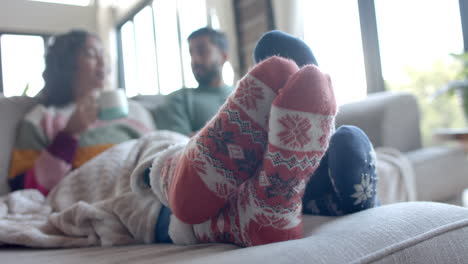 happy biracial couple on sofa in warm socks under blanket drinking coffee at home, slow motion