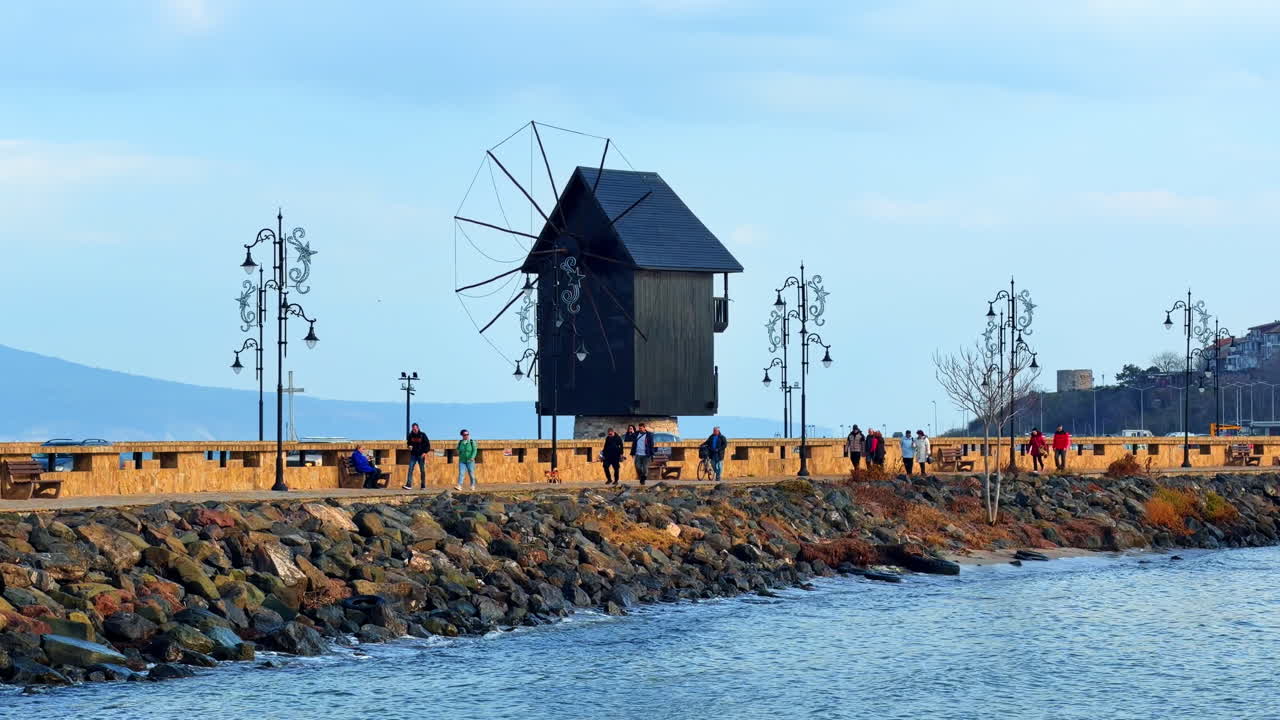 Burgas, Bulgarien, 29 oktober 2025: Människor promenerar längs vattnet vid väderkvarnen nära stranden i en lugn stad. Människor flanerar längs strandpromenaden nära en väderkvarn en klar dag vid havet.