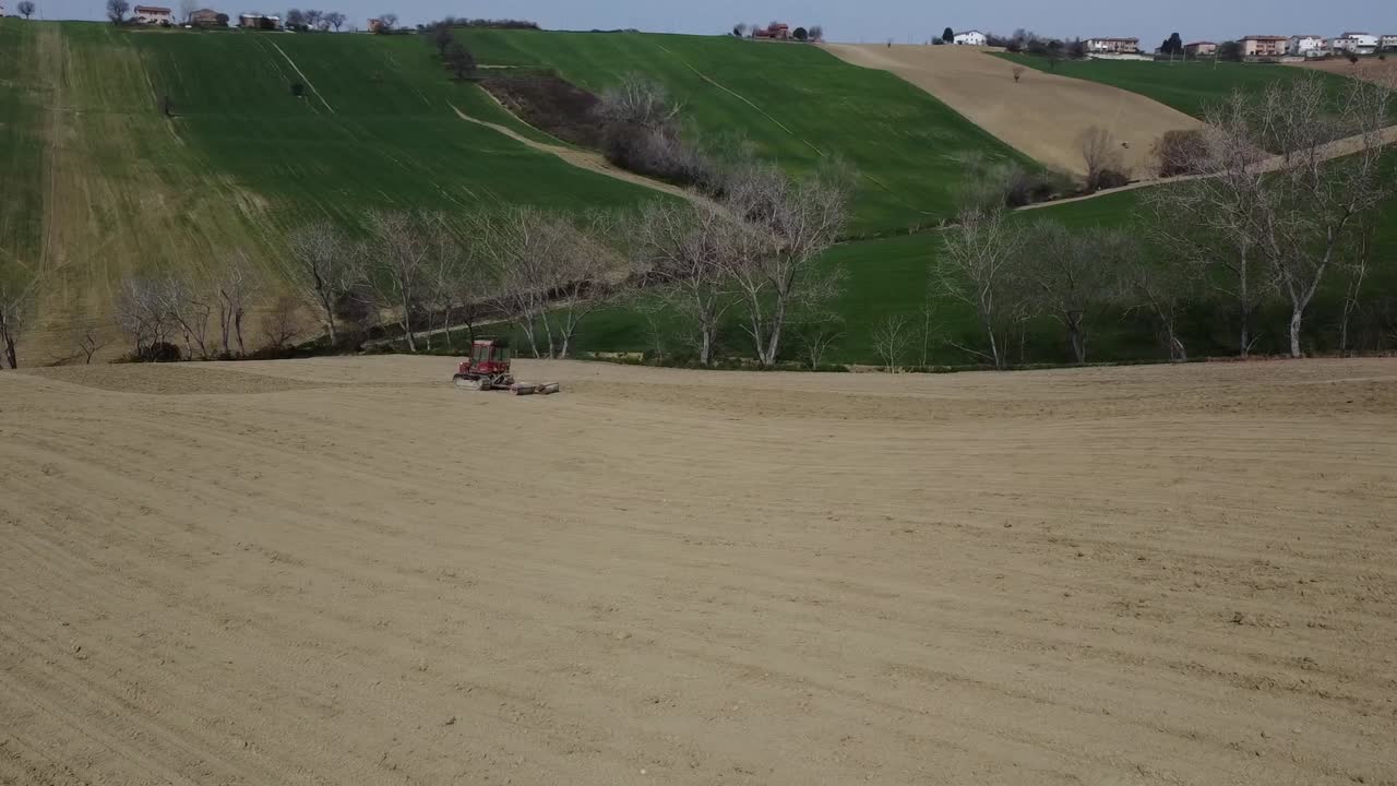 Drone rotating around red tractor soil rolling plowing land in scenic hill in Italian countryside, aerial farming agricultural job