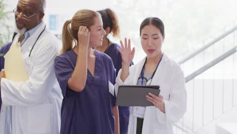 Video-of-two-diverse-female-doctors-looking-at-tablet-talking-in-busy-hospital-corridor