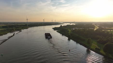 Ein-Frachtschiff-Fährt-Bei-Sonnenuntergang-Einen-Fluss-Hinunter,-Im-Hintergrund-Sind-Windturbinen-Zu-Sehen