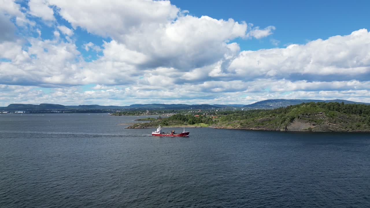 A red cargo ship moves through Oslo Fjord under a bright sky, with green islands and distant mountains in view