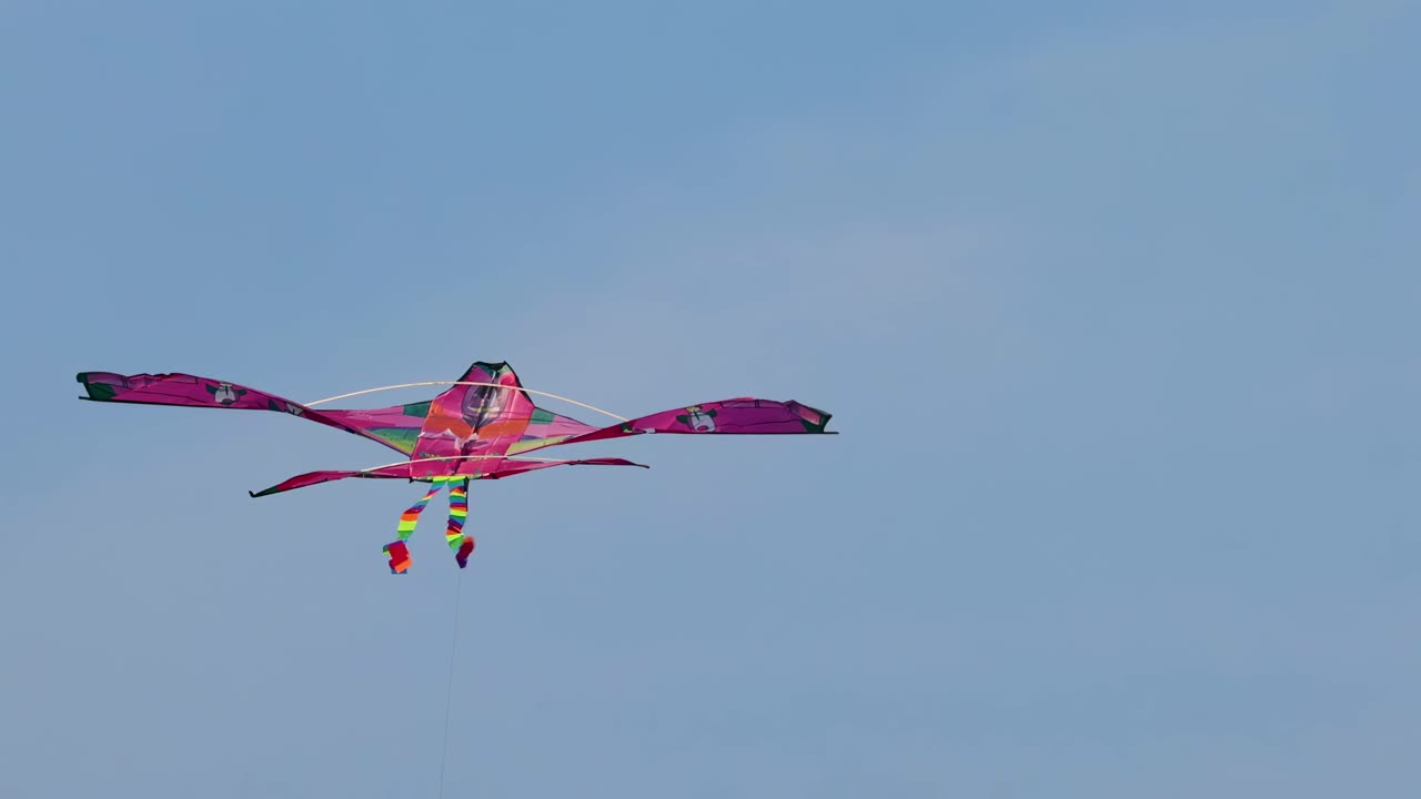 A colorful pink kite with a rainbow tail gracefully glides against a clear blue sky.