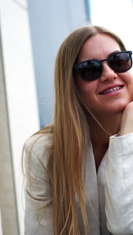 relaxed young happy woman sitting outdoors. blonde lady wearing white jacket and black sunglasses smiling to the camera. blurred backdrop. vertical video