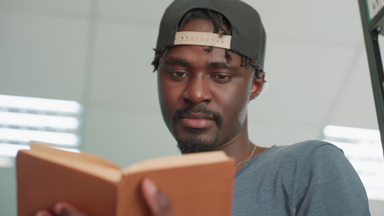 Primo piano di uno studente concentrato con un cappello che legge e sfoglia un libro con copertina rigida marrone in biblioteca, con luce intensa e libreria sullo sfondo