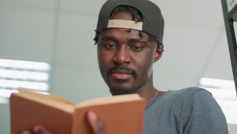 Close up of focused student wearing cap reading and flipping through brown hardcover in library, with bright light and bookshelf in background