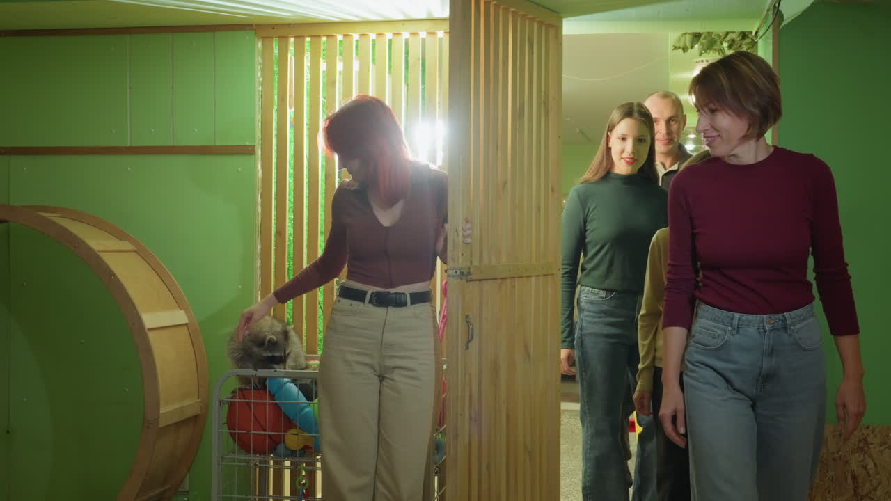green play wooden slats group entering light redheaded leader guides siblings and guardian through doorway toward colorful activity board, children examine balls and sensory toys while teacher greets