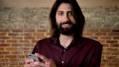 joven atractivo hombre moreno con barba está viendo en el teléfono inteligente, viendo en la cámara, fondo de ladrillo, concepto de comunicación