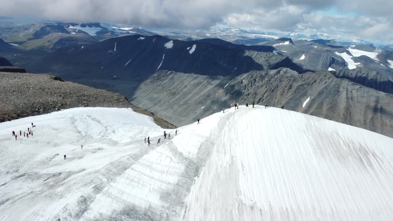 Aerial drone view ascending and flying up showing a group of people hiking and walking on a shite snow covered hill or a mountain in Sweden during a cloudy day while large Kebnekaise mountains visible