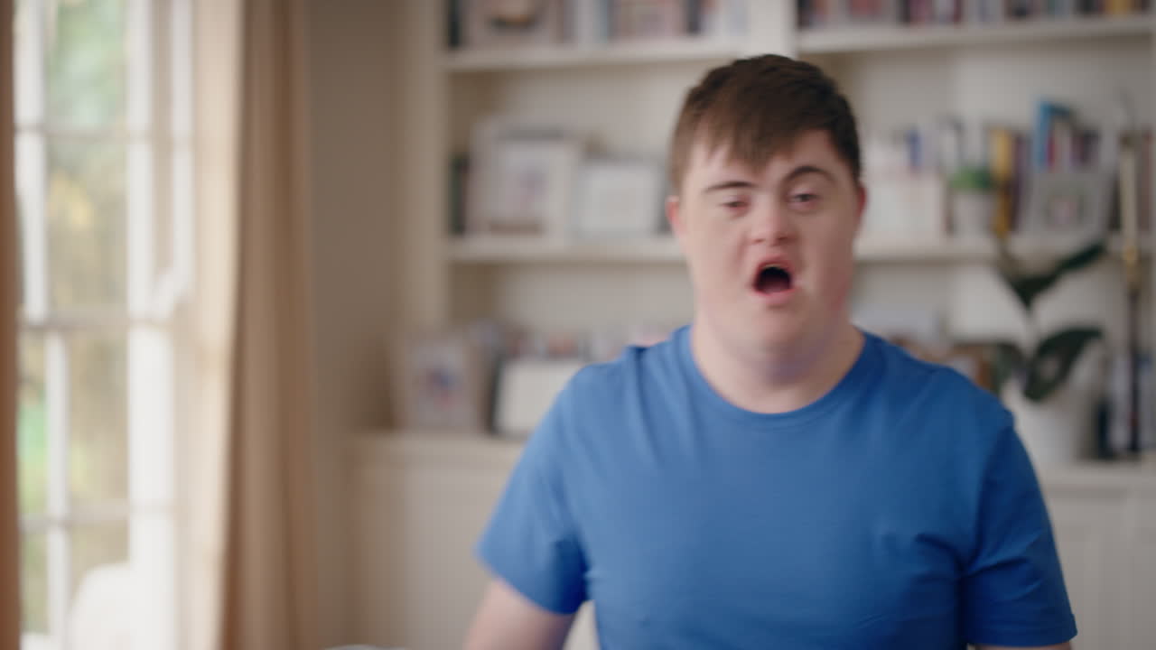 다운 증후군을 가진 재미있는 십대 소년이 거실에서 춤을 추고 특별한 요구를 가진 아이가 재미를 느끼고 어리석은 춤 움직임으로 축하하고 집에서 행복한 주말을 즐기고 4k