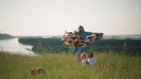 una mujer con una túnica azul, sostiene una bufanda a cuadros mientras el viento la sopla suavemente en un campo cubierto de hierba con vistas a un río pintoresco. dos niños felices con camisas blancas se sientan a su lado, inclinándose