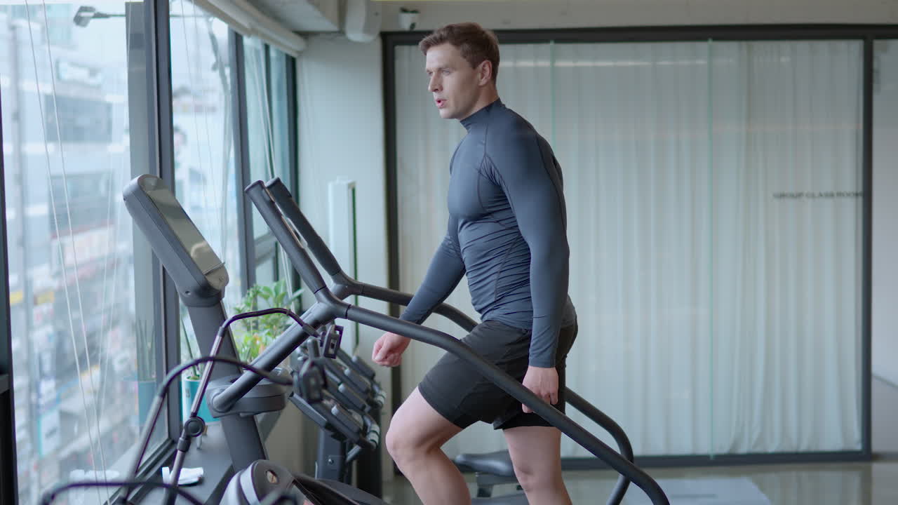 Athletic Man Exercises on Stepmill Stair Climber Machine in Modern Gym Looking Though Window On City Street in slow motion