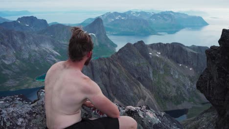 shirtless man sitting on mountain peak of kvaenan overlooking mountains and fjord in senja, norway