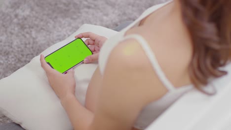 mano cercana de una mujer embarazada sosteniendo un teléfono móvil con pantalla verde o llave cromada sentada en la alfombra en casa. vista superior mujer embarazada usando un teléfono inteligente con pantalla verde para comprar en la tienda de bebés en línea
