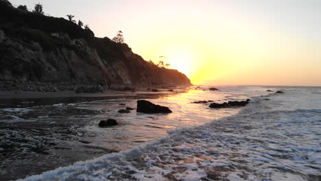 A-beach-at-sunrise-with-ocean-waves-and-reflections-on-the-shore-and-cliffs-in-silhouette-along-the-Santa-Barbara,-California-coast-AERIAL-DRONE