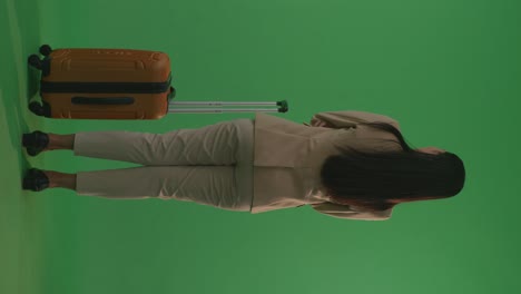 full body of back view asian businesswoman traveler with luggage crossing arms and looking around while standing in the green screen background studio, during sunset time