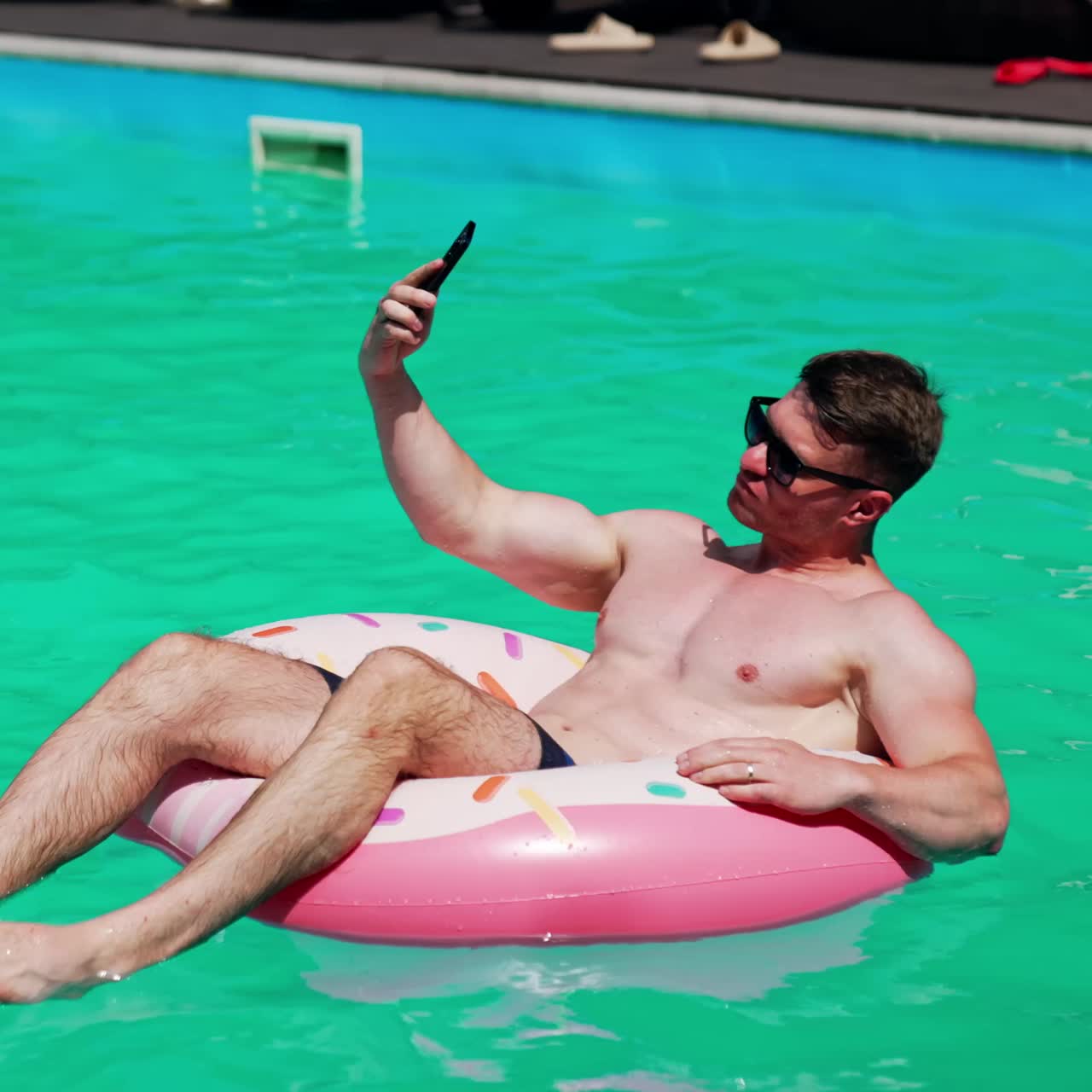 Relaxed Caucasian sportive man swimming in the pool. Man sits in the inflatable donut having a video chat on his phone