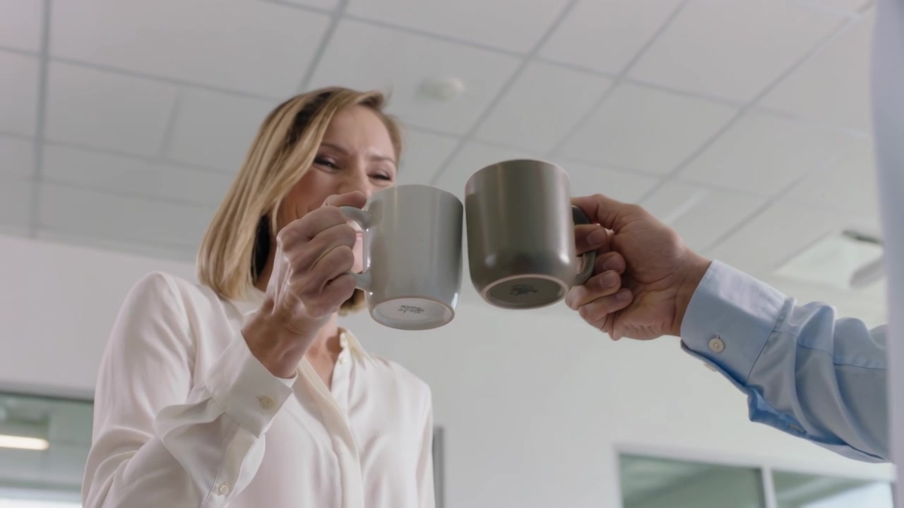Mulher sorridente de blusa clara levantando uma caneca branca enquanto um colega estende uma caneca cinza no escritório, brindando.