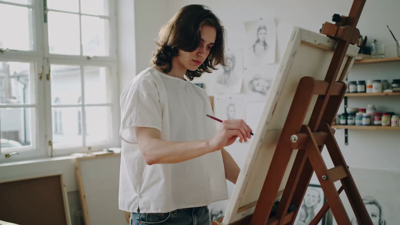 Painting artist lifting brush, marking at studio, refining work on easel in white top, jeans