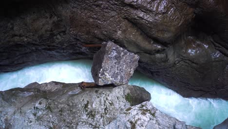 Ein-Blauer,-Kalter-Und-Klarer-Gebirgsfluss-Fließt-Durch-Eine-Schlucht-In-Den-Alpen,-Wasser-Schäumt-Auf,-Im-Vordergrund-Ein-Großer-Stein