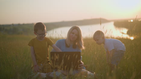 a woman in a blue gown sits on a scarf in a grassy field, working on a laptop during sunset. beside her, a young boy dressed in yellow sits, while another boy in white bends beside them