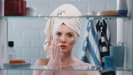 View-Through-Bathroom-Cabinet-Of-Woman-Wearing-Towel-On-Her-Head-Putting-On-Highlighter-Make-Up