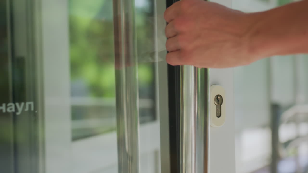 White Man Disinfecting Glass Door Handle With Wipe, Messenger Bag Strap Visible, Sunlit Porch Reflection, Repetitive Motion Suggesting Compulsive Cleanliness And Careful Hygiene Routine