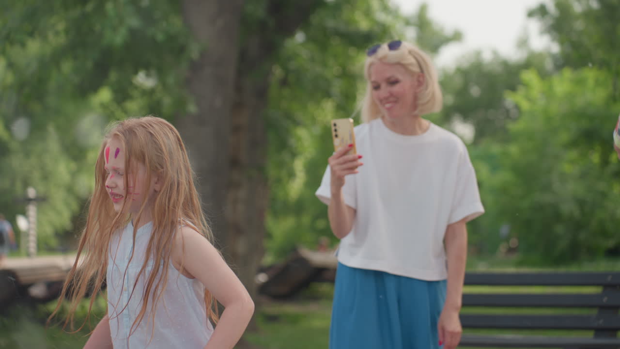 cheerful mother in casual wear gives daughter kiss while filming playful moment with phone in green park, smiling girl with long hair, outdoor family bonding captured on summer day