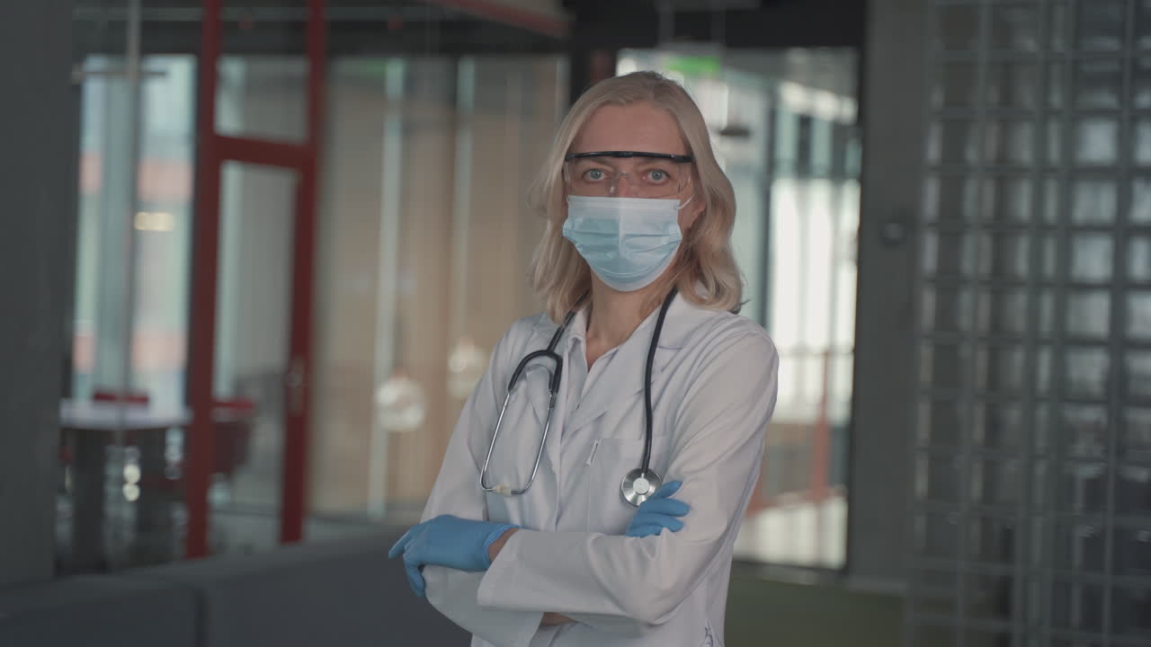A Blonde, Middle Aged Female Doctor In A Facemask Looks Directly At The Camera With Crossed Arms