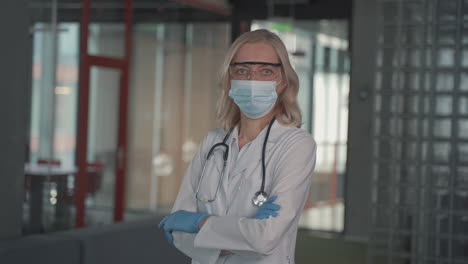 a blonde, middle aged female doctor in a facemask looks directly at the camera with crossed arms