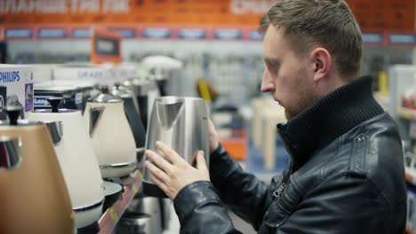 young man choosing good electric kettle for his new house in a big shop with is a big choice of small household appliances