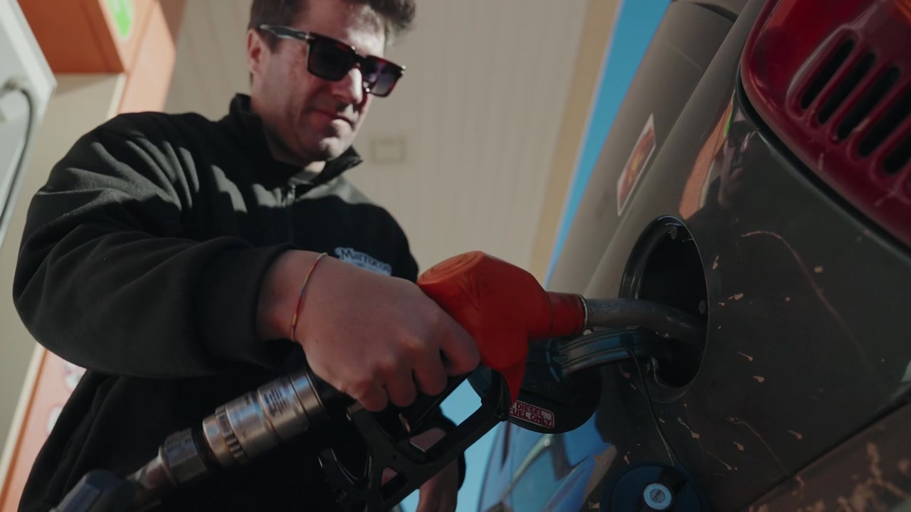 Mand med solbriller trækker rød tankpistol ud af bilens tankstuds efter tankning på en tankstation