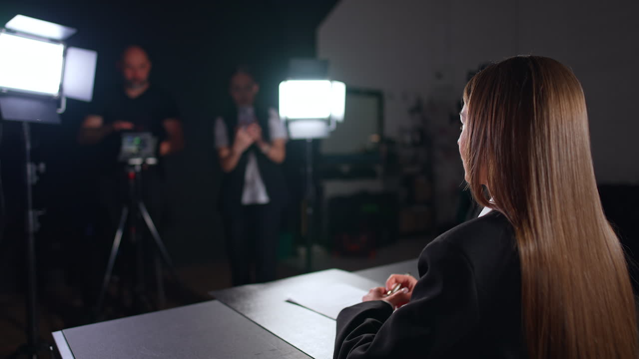 Brunette woman in black jacket sits at desk talking to camera. Filming crew works backstage. Video recorded in studio.