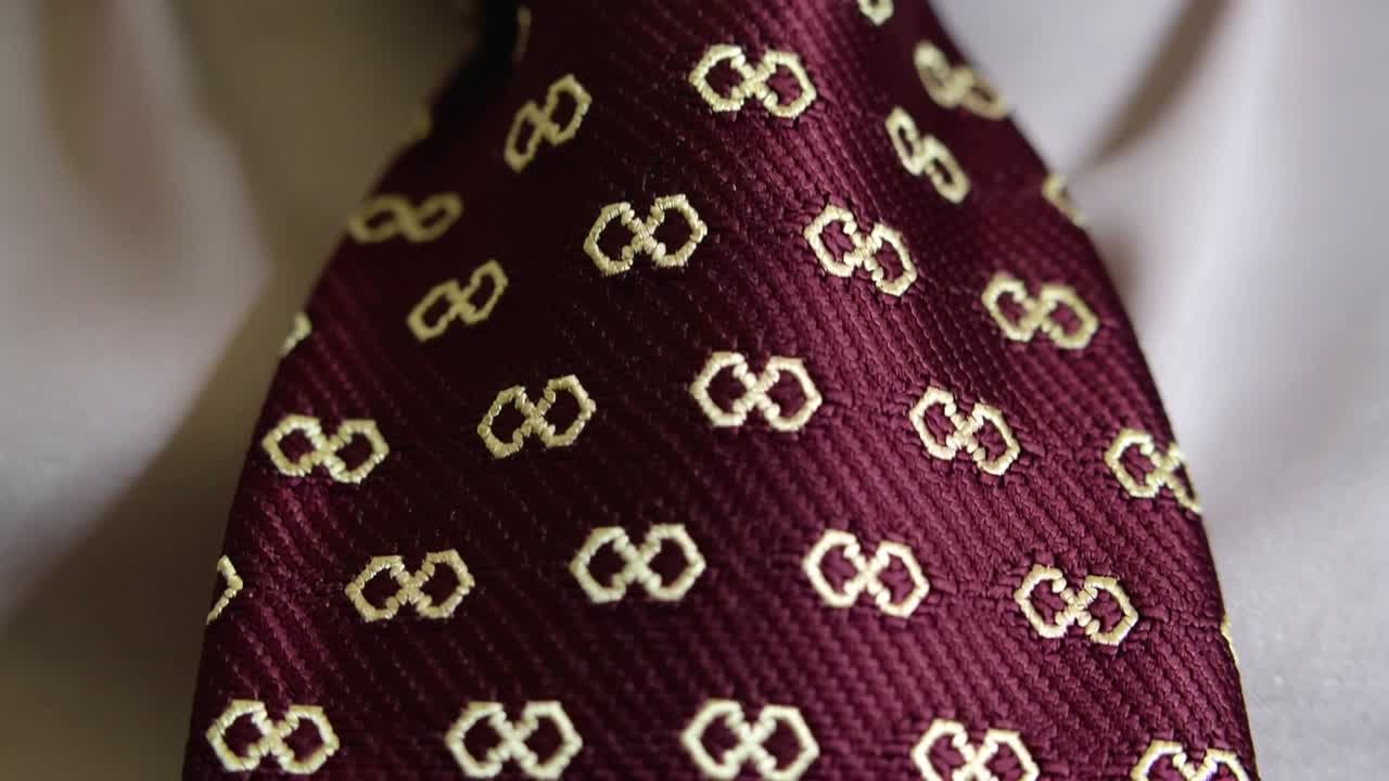 Una telecamera che ingrandisce rivelando un primo piano del nodo e della pala di una cravatta bordeaux, evidenziando un motivo dorato e il colletto della camicia.
