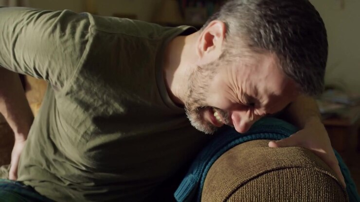 Man in olive shirt sitting on a living room sofa grimacing, leaning over the armrest holding his lower back