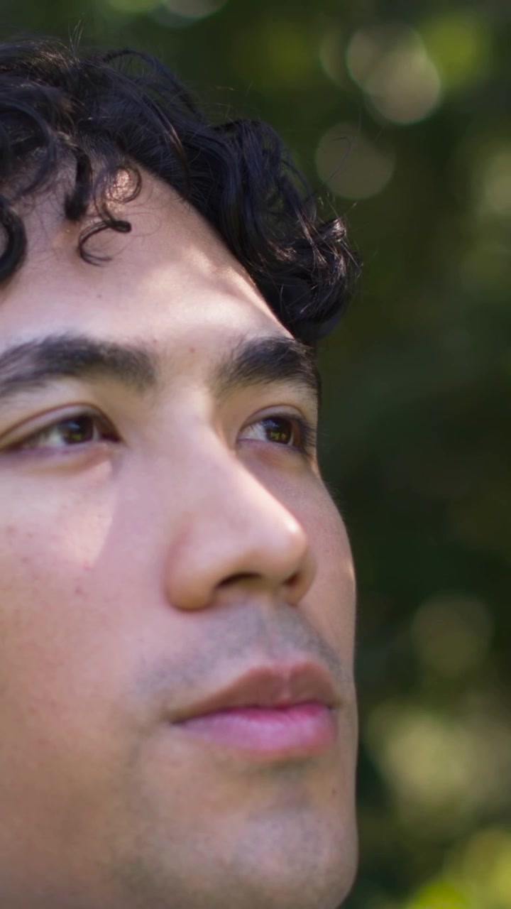 Video verticale: Uomo in camicia che apre gli occhi e inclina la testa all'insù in un parco, mentre il sole a chiazze crea un bokeh fogliare in movimento.