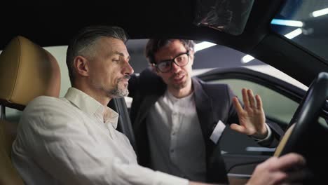 un hombre confiado de mediana edad con cabello gris y una camisa blanca se comunica con un asistente de concesionario de automóviles en un traje de negocios mientras evalúa el interior del coche desde adentro