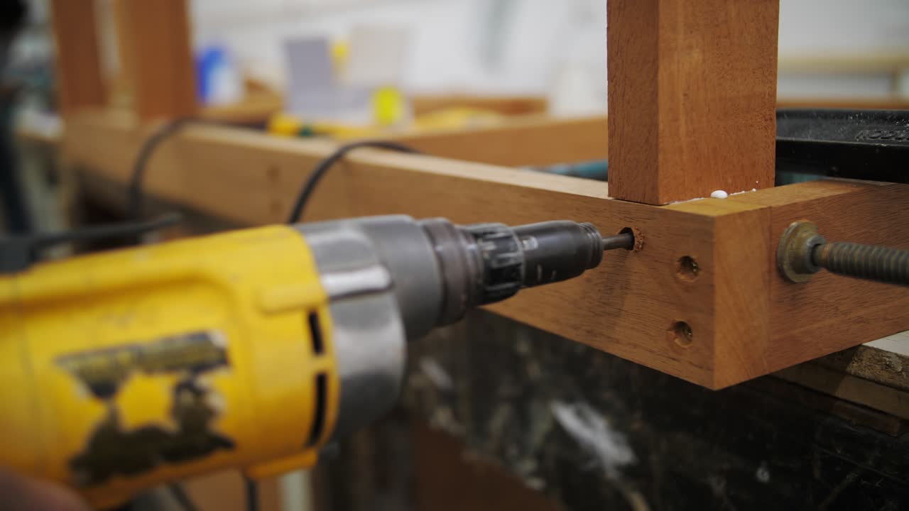 The image shows a yellow power drill being used to drive a screw into a wooden frame. The wood is clamped securely, and the background features a workshop environment with tools and materials visible.