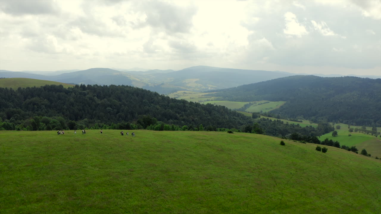 Group of tourists walking across vast green meadow surrounded by forested hills and misty mountains under cloudy summer sky. Aerial establishing shot. Bieszczady, Poland