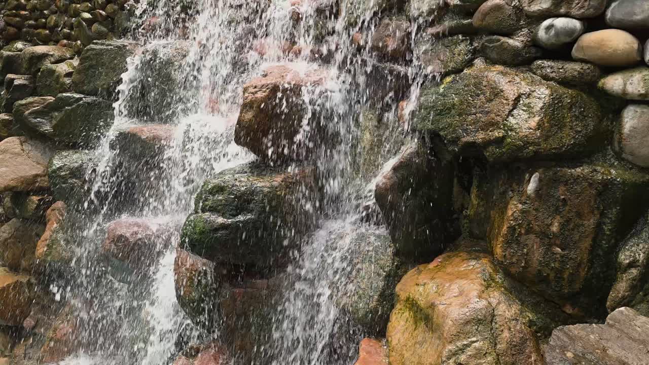 A close-up shot captures the details of a beautiful garden waterfall, with clear water cascading and splashing over dark, moss-covered rocks in a peaceful and relaxing nature scene