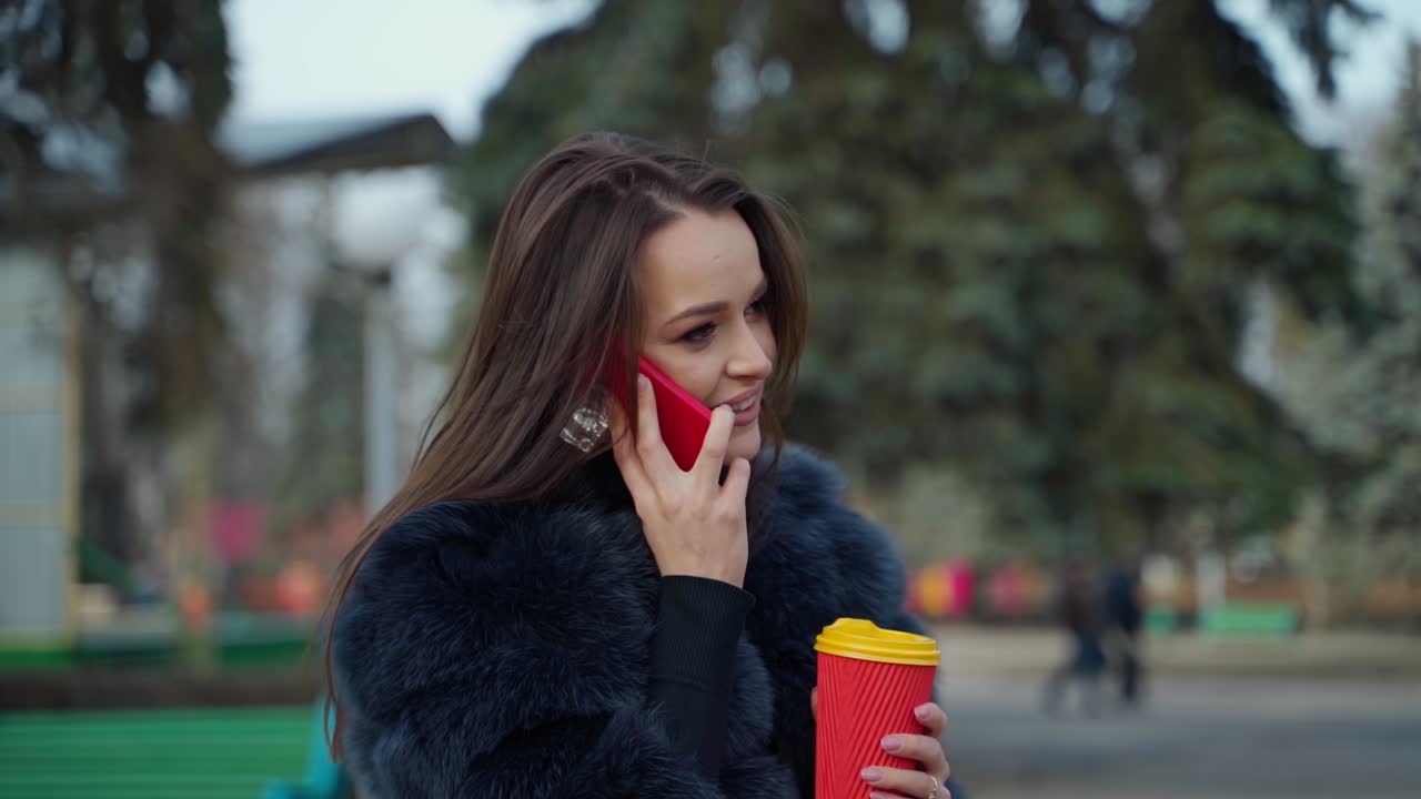 Charming girl in fluffy coat. Beautiful young brunette talk the mobile phone and smiles on blur park background. Woman in fur coat holds plastic cup of coffee outdoors.