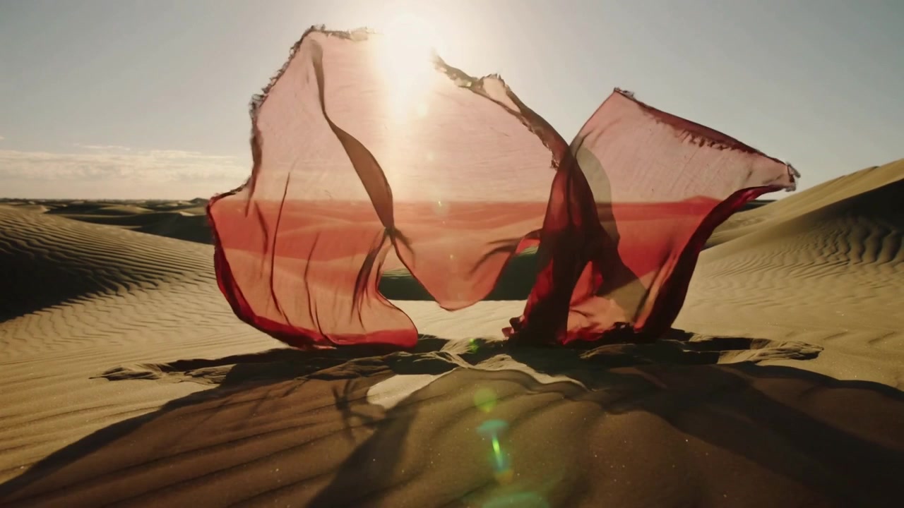 Vento levantando tecido vermelho, dançarina se movendo sob ele em duna, criando formas em camadas iluminadas pelo sol