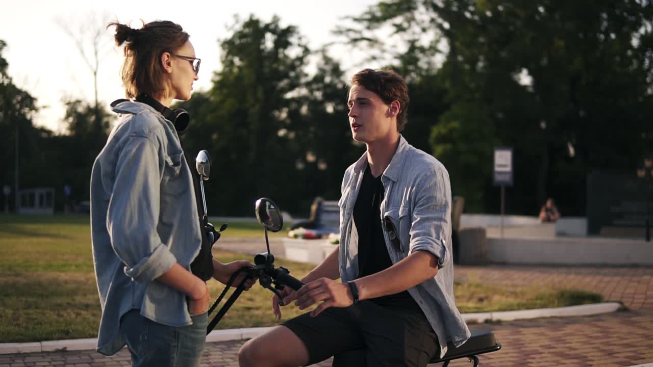 una pareja joven con las mismas camisas azules en la calle. un niño está sentado en una bicicleta eléctrica, gesticulando y hablando con su novia frente a él. estilo de vida feliz. cámara lenta