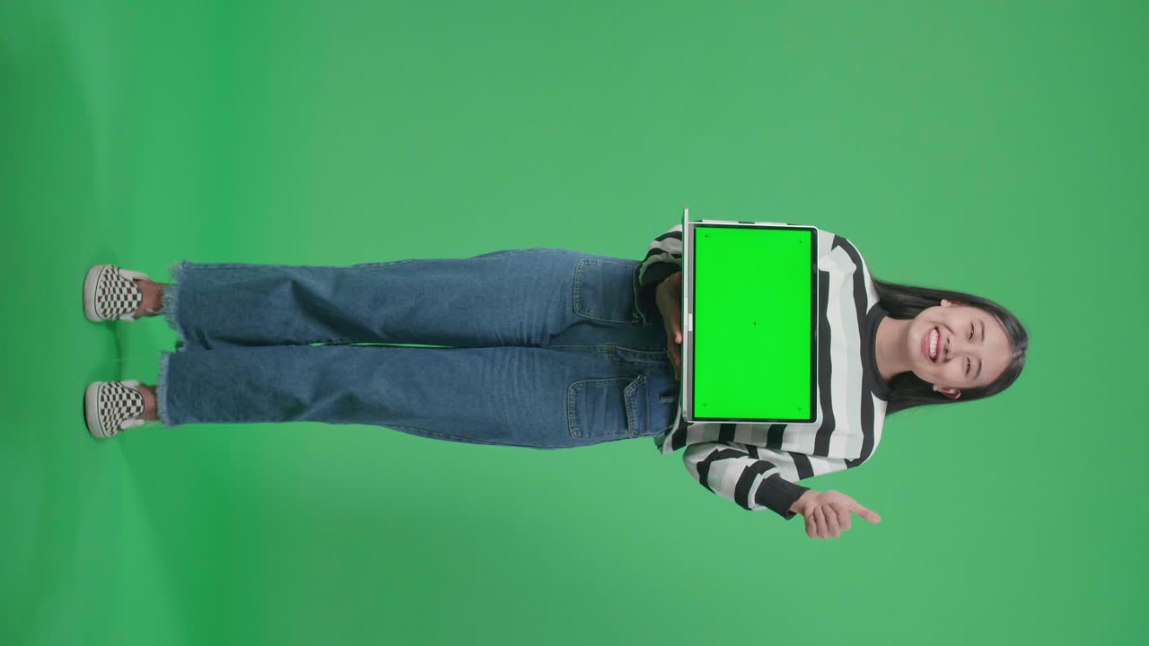 Full Body Of A Smiling Asian Woman Showing Thumbs Up Gesture While Holding Green Screen Computer In The Green Screen Studio