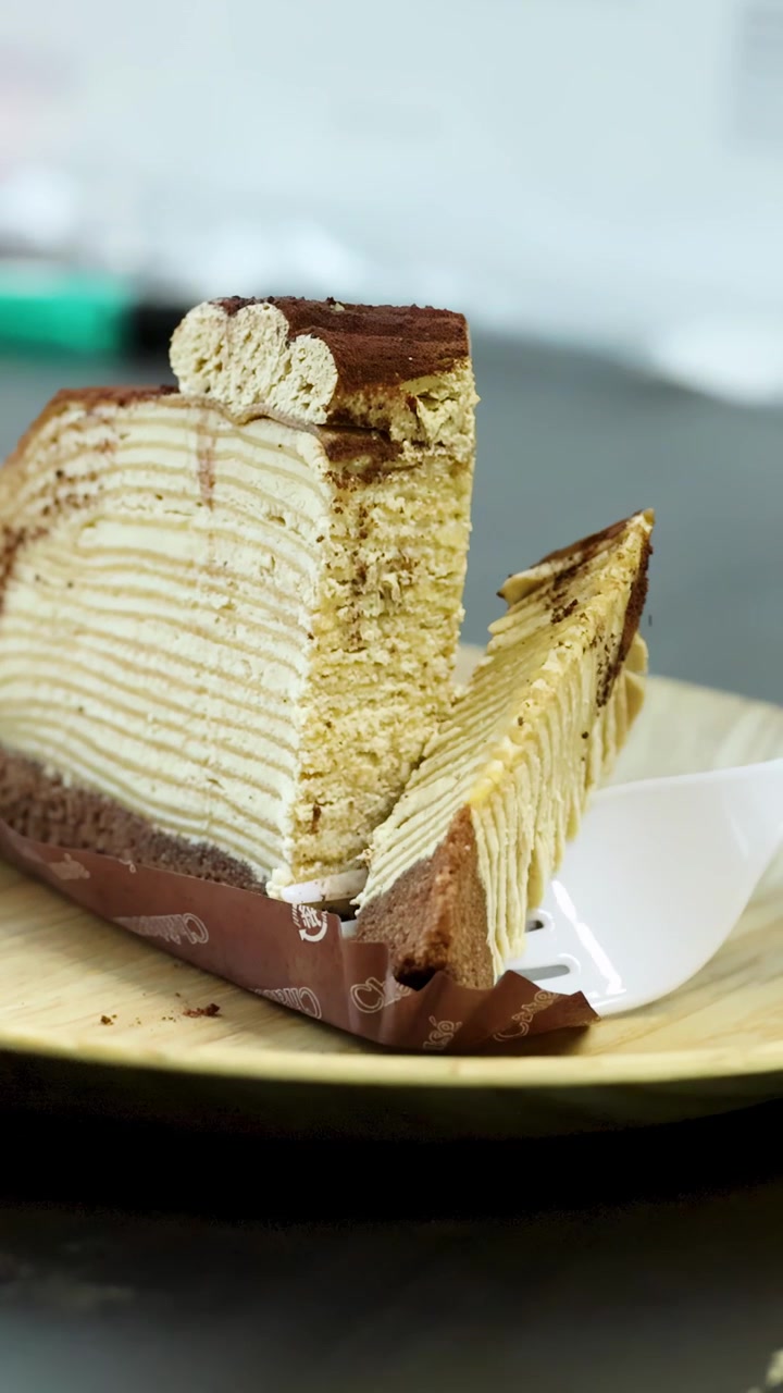 Una forchetta di plastica taglia e solleva una torta di crepes al tiramisù a strati su un piatto di legno, con illuminazione interna soffusa.