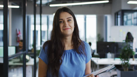 Portrait-of-caucasian-businesswoman-standing-in-office-holding-paperwork-smiling-to-camera