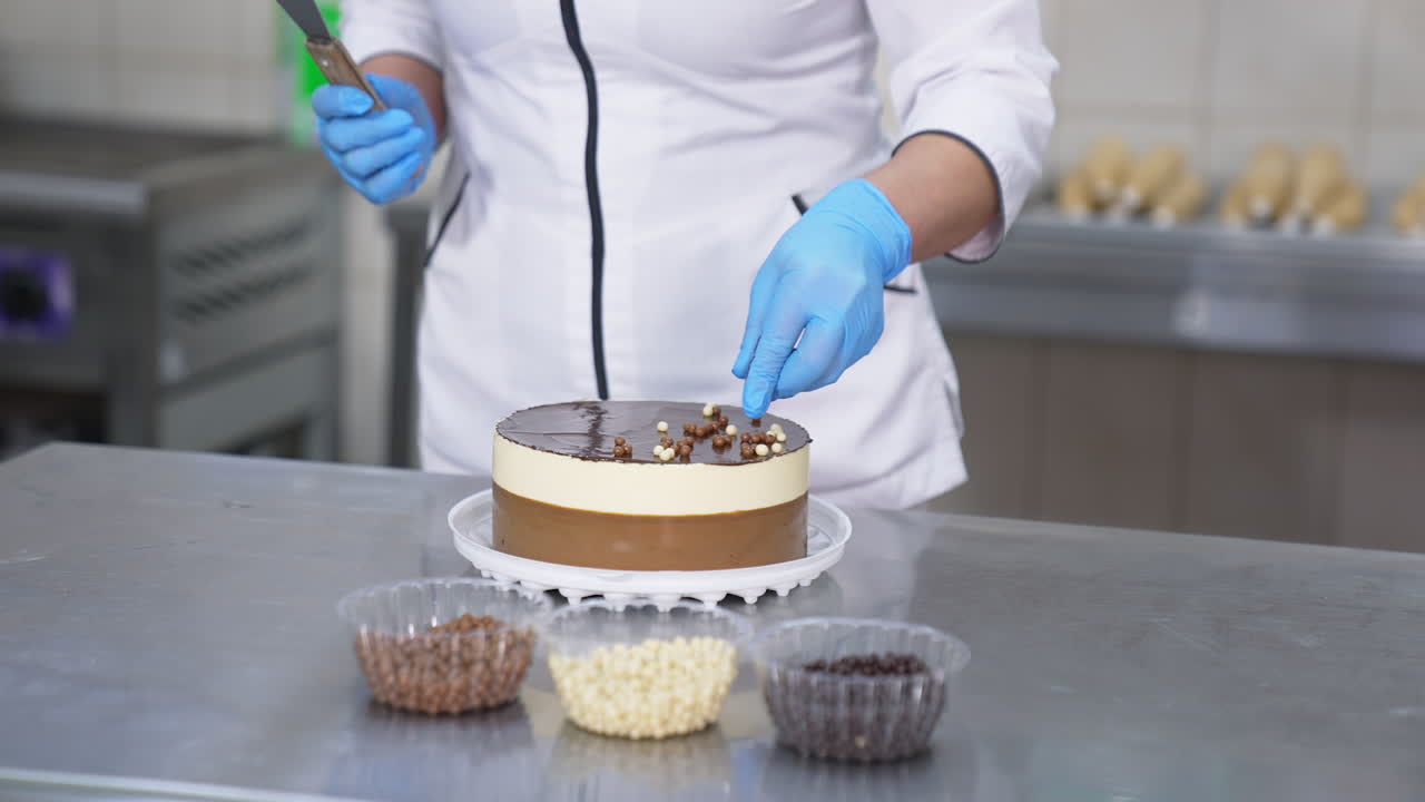 White and chocolate cake being decorated by the confectioner. Woman in gloves takes different candies from plastic cups and sprinkles them on top of the cake.