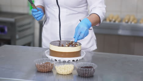 white and chocolate cake being decorated by the confectioner. woman in gloves takes different candies from plastic cups and sprinkles them on top of the cake.