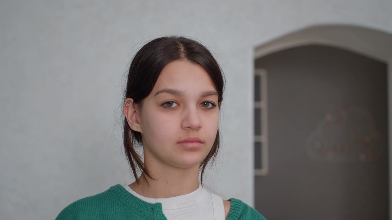 Giovane donna in un appartamento vuoto sta in silenzio tra pareti spoglie e un arco, che indossa un maglione verde, ritratto ravvicinato con luce diurna soffusa, espressione sottilmente introspettiva durante il disimballaggio e l'insediamento