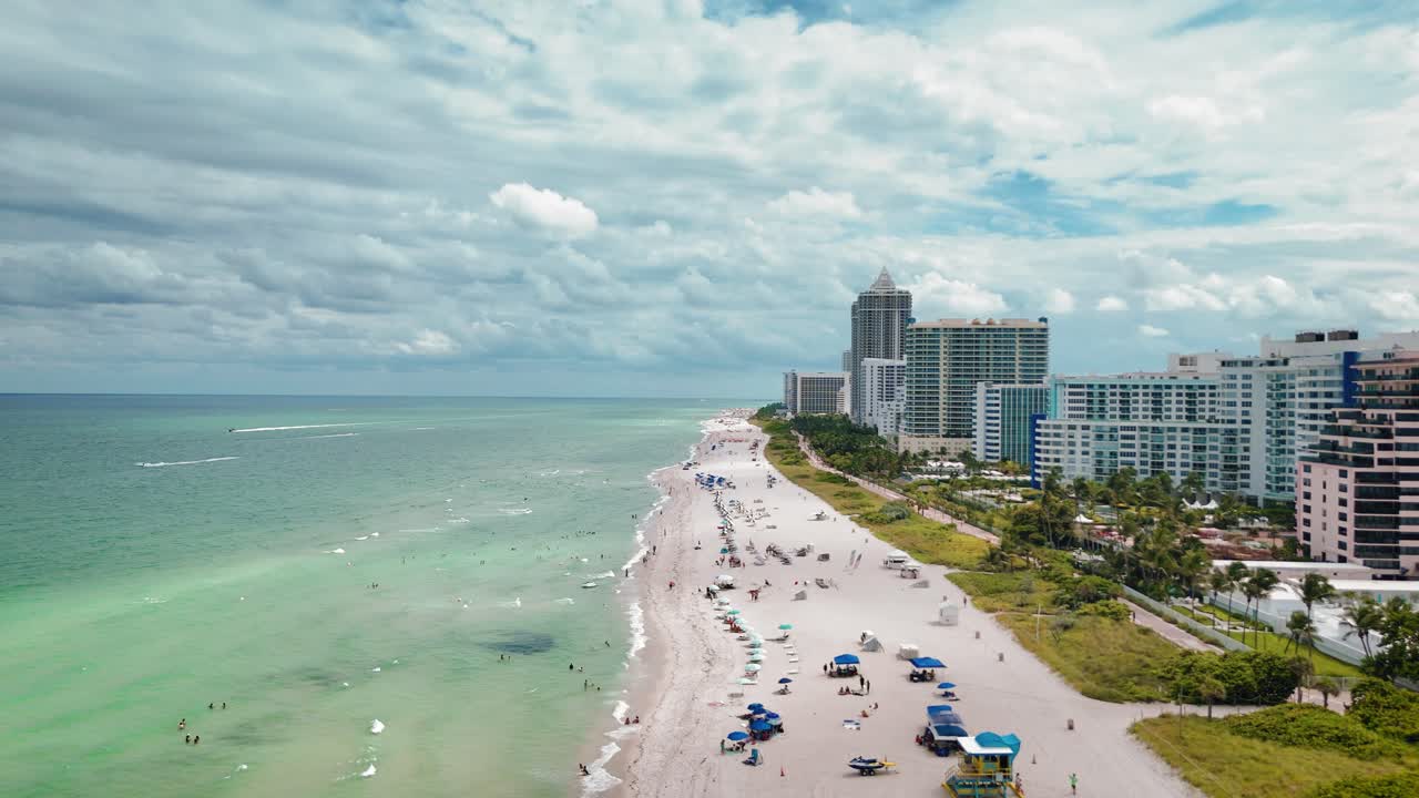 Breathtaking drone panorama of Millionaire's Row in Miami Beach. High-rise buildings line a crowded beach next to turquoise ocean waters. Perfect for urban, travel, and commercial real estate themes